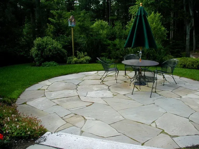 A circular flagstone patio with a round metal table, four chairs, and a dark green umbrella in a lush, grassy garden.