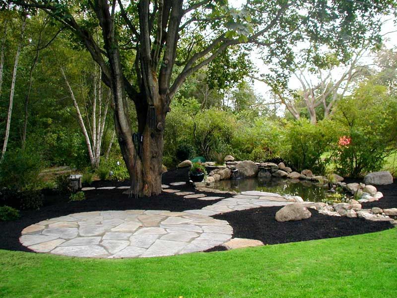 A stone patio and a rock-bordered pond sit in a garden landscape with a large central tree, green foliage, and black mulch.