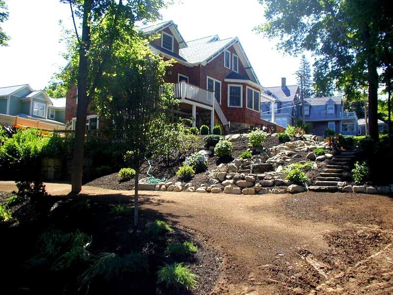 A rustic brown house set on a sloped, landscaped lot with stone retaining walls, rocky garden beds, and a gravel driveway.