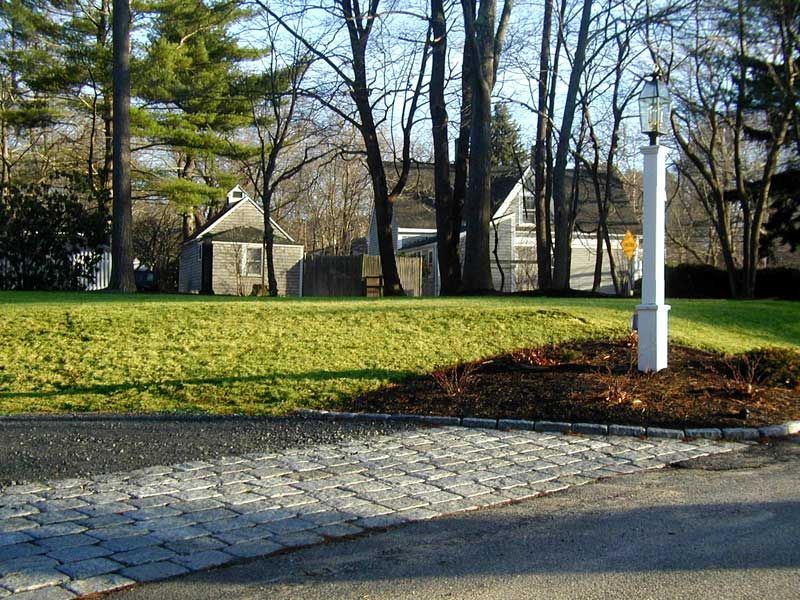 A white lantern post stands on a lawn next to a cobble-bordered driveway leading toward a house and trees.