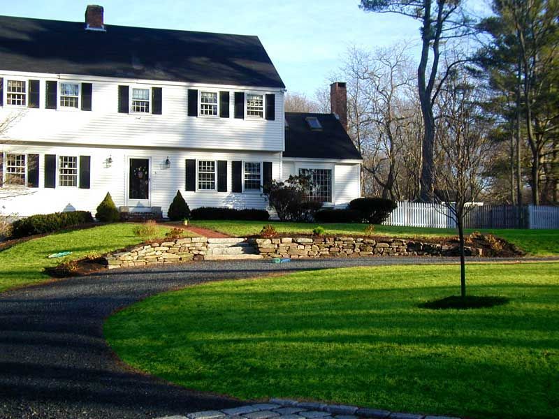 A two-story white colonial house with black shutters, a stone retaining wall, and a curved gravel driveway in a yard.