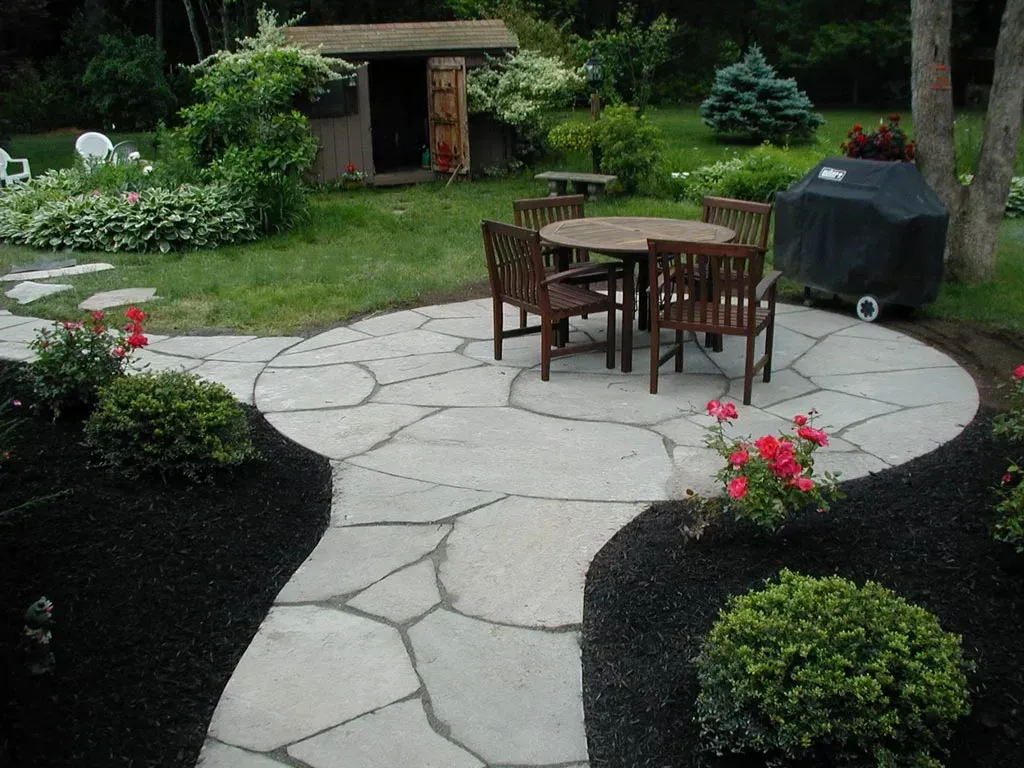 A stone patio with a round dining table and chairs, surrounded by garden beds with flowers and shrubs in a backyard.