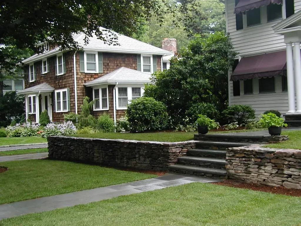 A stone retaining wall with steps leads to a white house, with a shingled home and garden in the background.