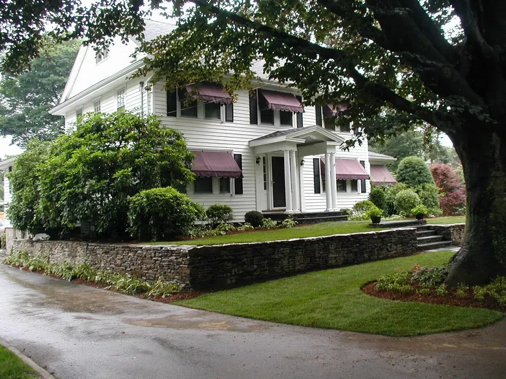 A white two-story house with dark shutters and maroon awnings, set behind a stone retaining wall and large trees.