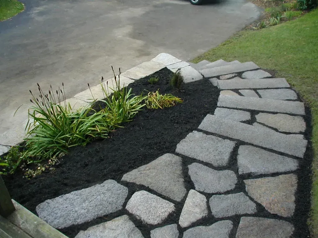 A curved stone pathway lined with dark mulch and green plants leading toward a driveway and grass lawn.