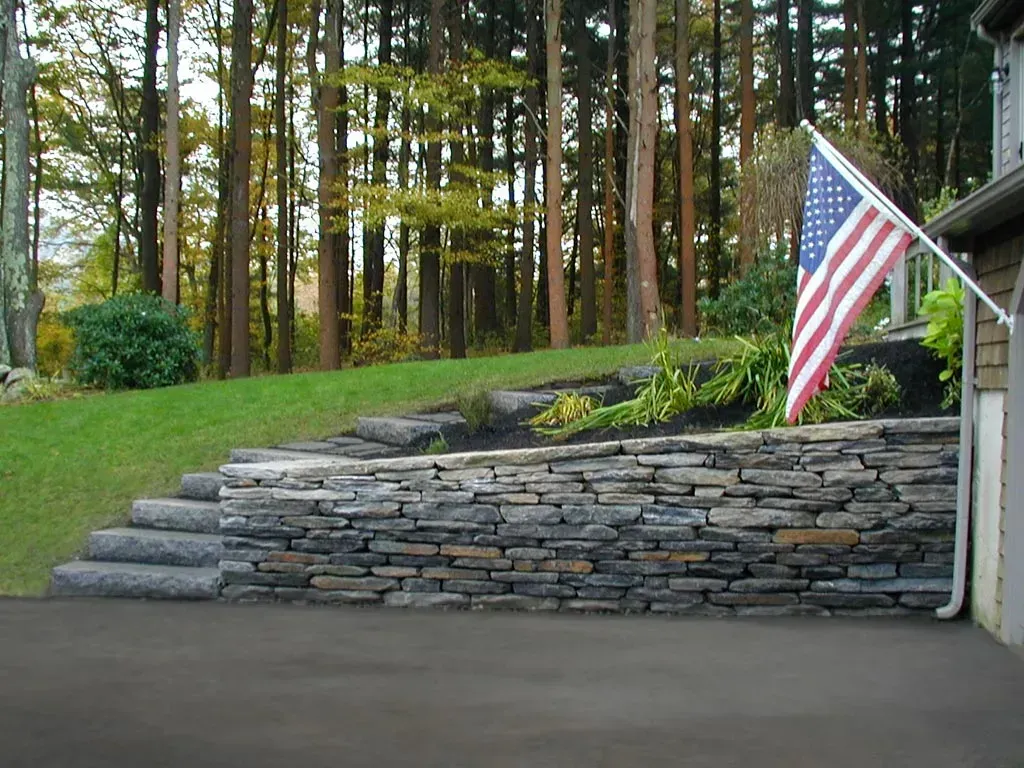 A stone retaining wall with integrated steps next to an asphalt driveway and a house with a flag, backed by forest trees.