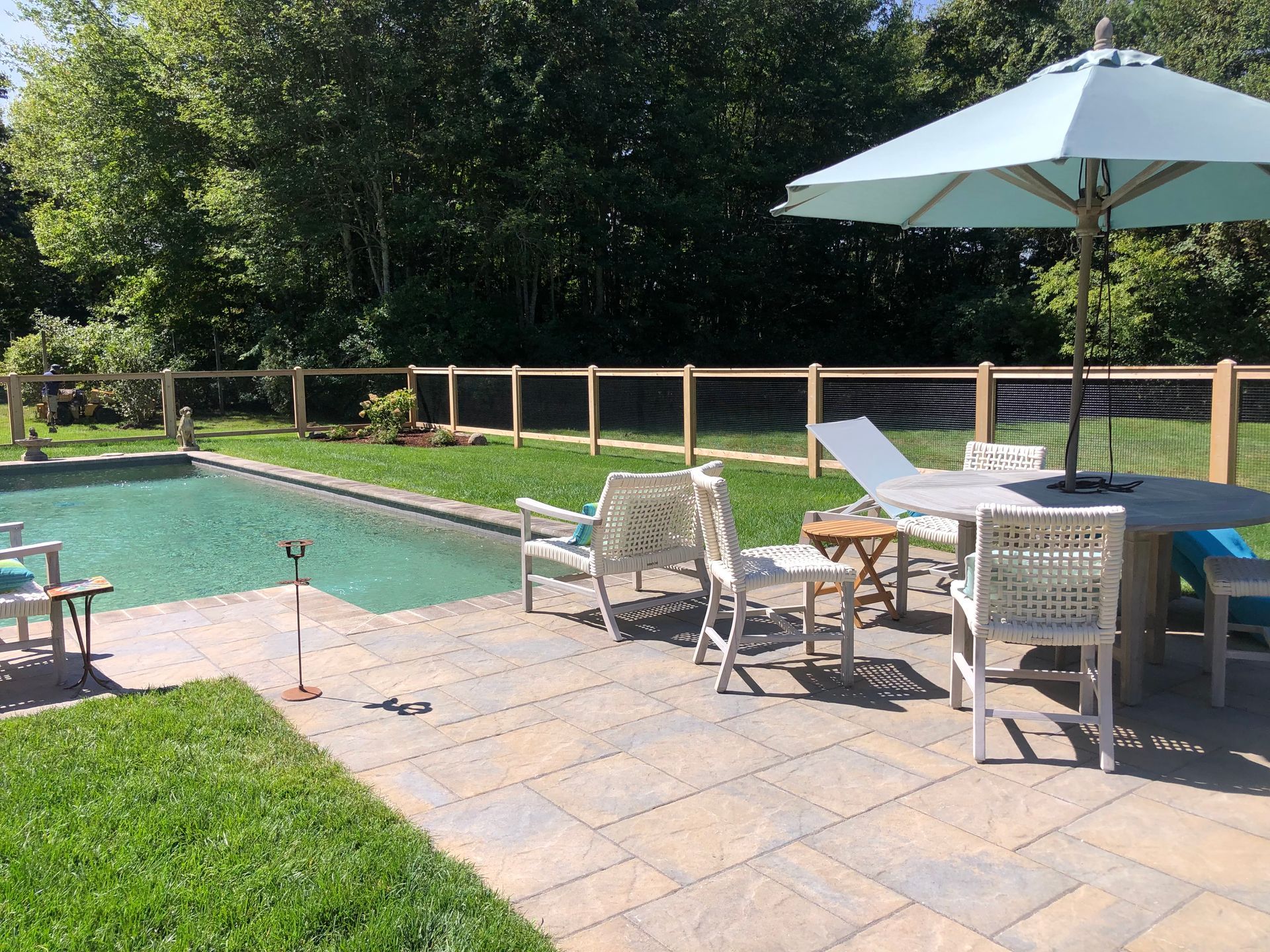 A backyard swimming pool with a stone patio, outdoor dining set, umbrella, and lounge chairs surrounded by a wooden fence.