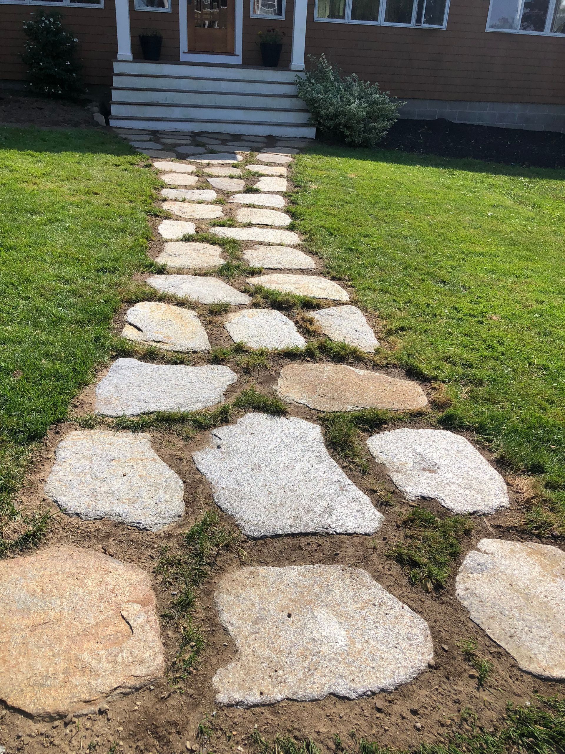 A stone flagstone walkway leads through a grassy front lawn to a set of stairs at the entrance of a brick house.