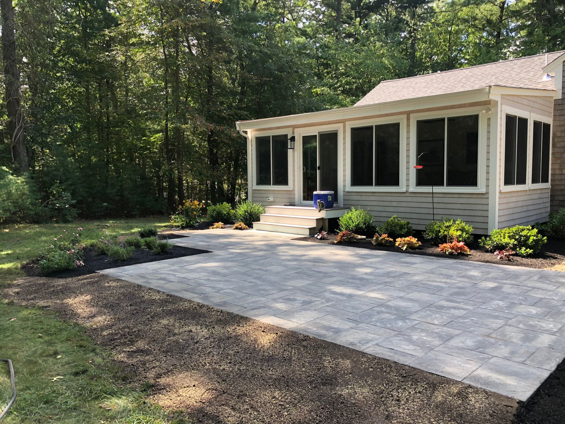 A light gray sunroom with a patio of light-colored pavers, surrounded by mulch beds and lush greenery in a forest setting.