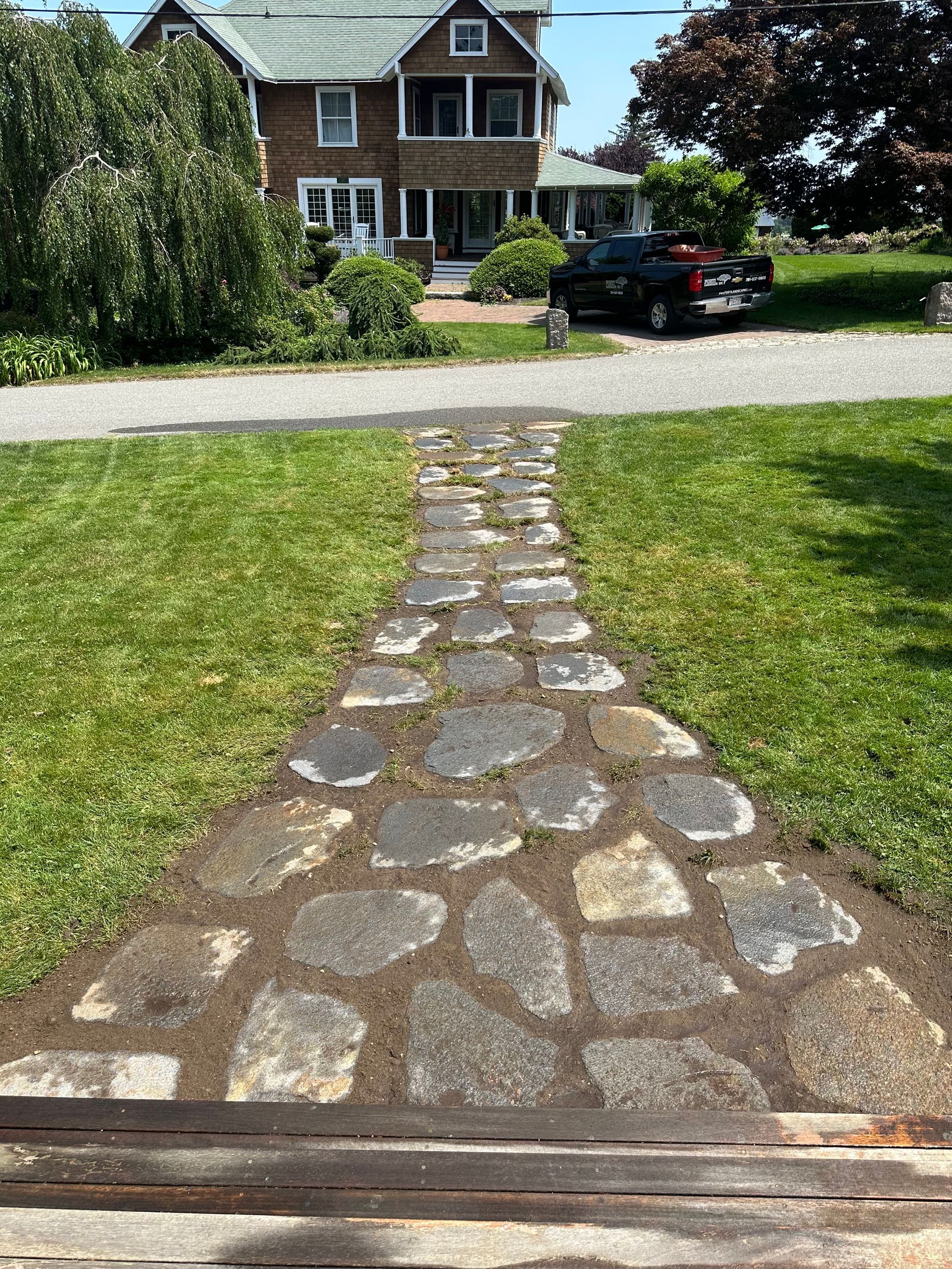A stone walkway leading through a grassy front yard toward a brown two-story house with a black pickup truck parked nearby.