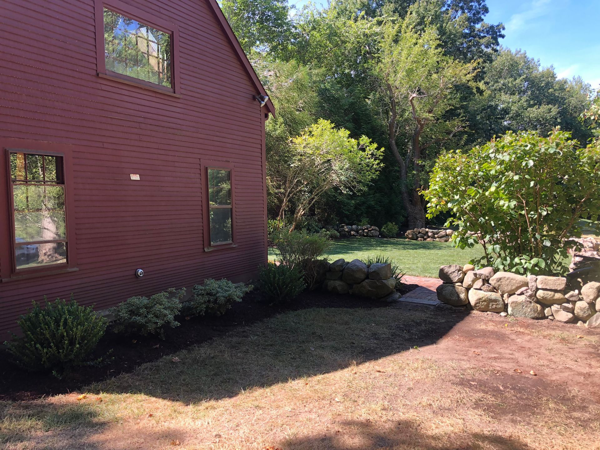 A red house exterior featuring a stone wall, manicured bushes, and a green yard under a clear blue sky.