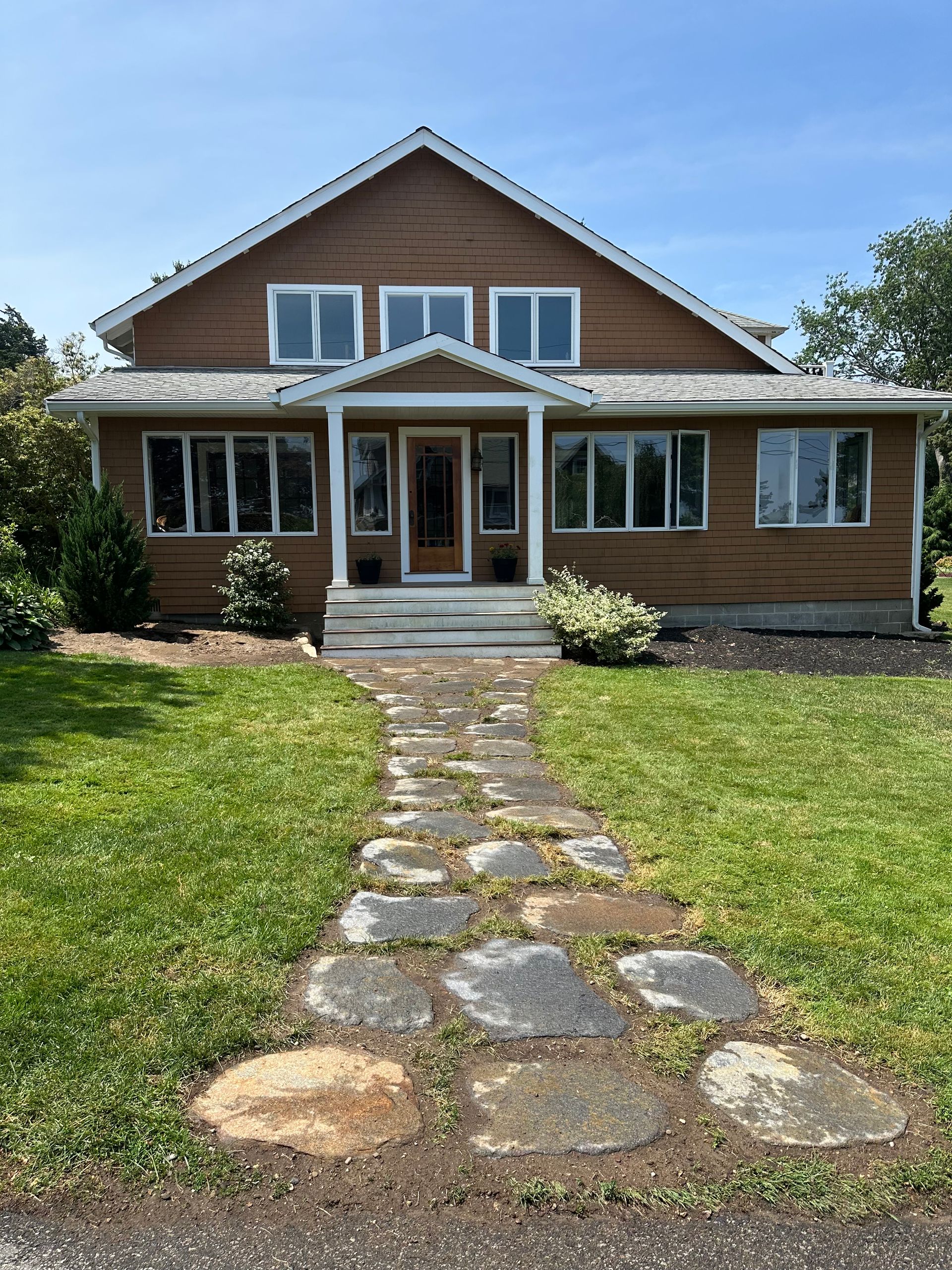A brown two-story house with a gabled roof and a stone walkway leading to a central front porch under a sunny blue sky.