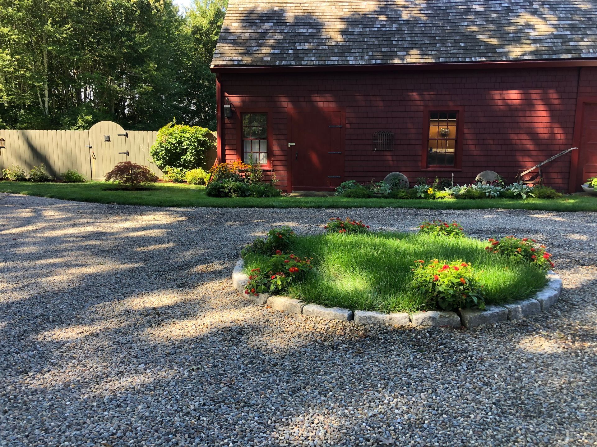 A red barn stands behind a stone-bordered circular garden bed filled with green grass and flowers in a gravel driveway.