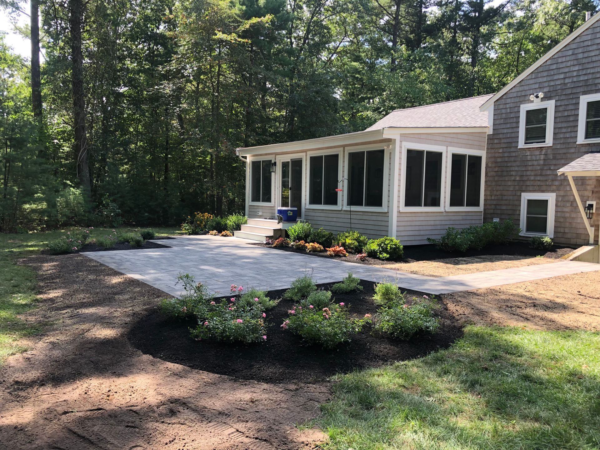 A light-colored stone patio leads to a screened-in porch with a gray shingle house, surrounded by mulched garden beds.