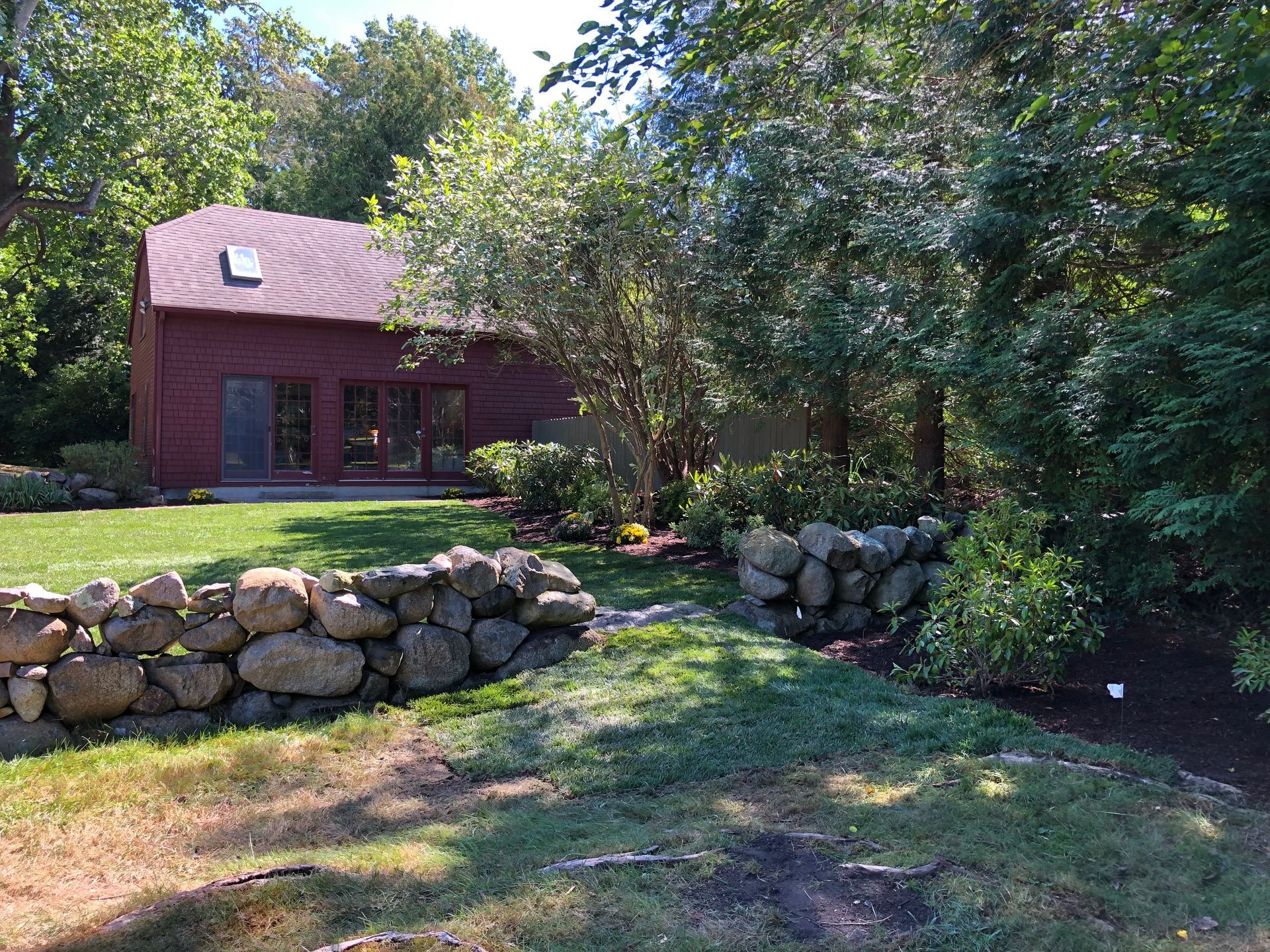 A red building sits in a wooded backyard behind a stone wall and a grassy lawn on a sunny day.