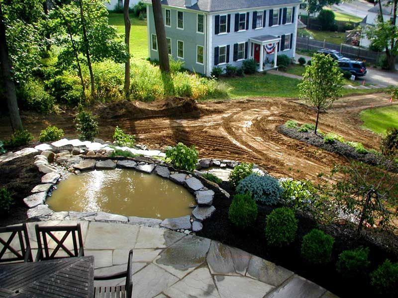 A stone patio with a small pond overlooks a backyard under construction and a gray house in the distance.