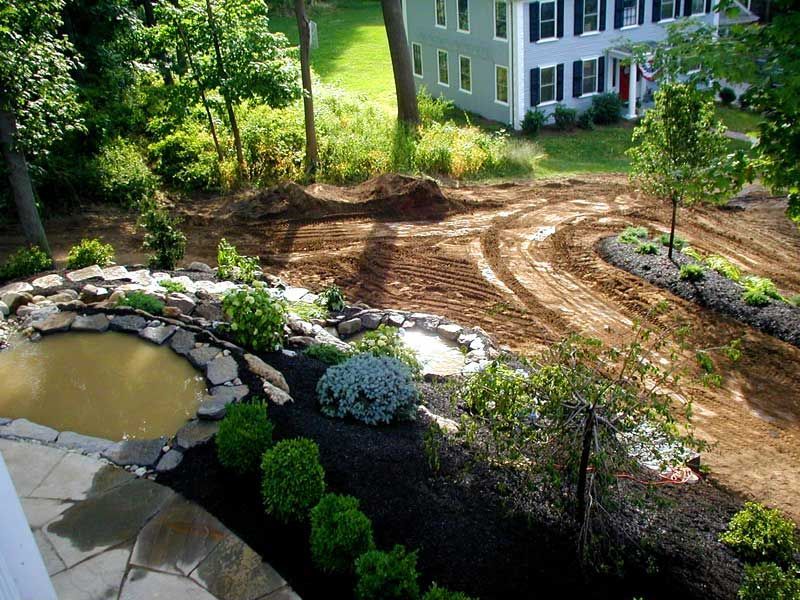 A pond with stone edging overlooks a recently cleared, muddy landscape leading to a two-story gray house.