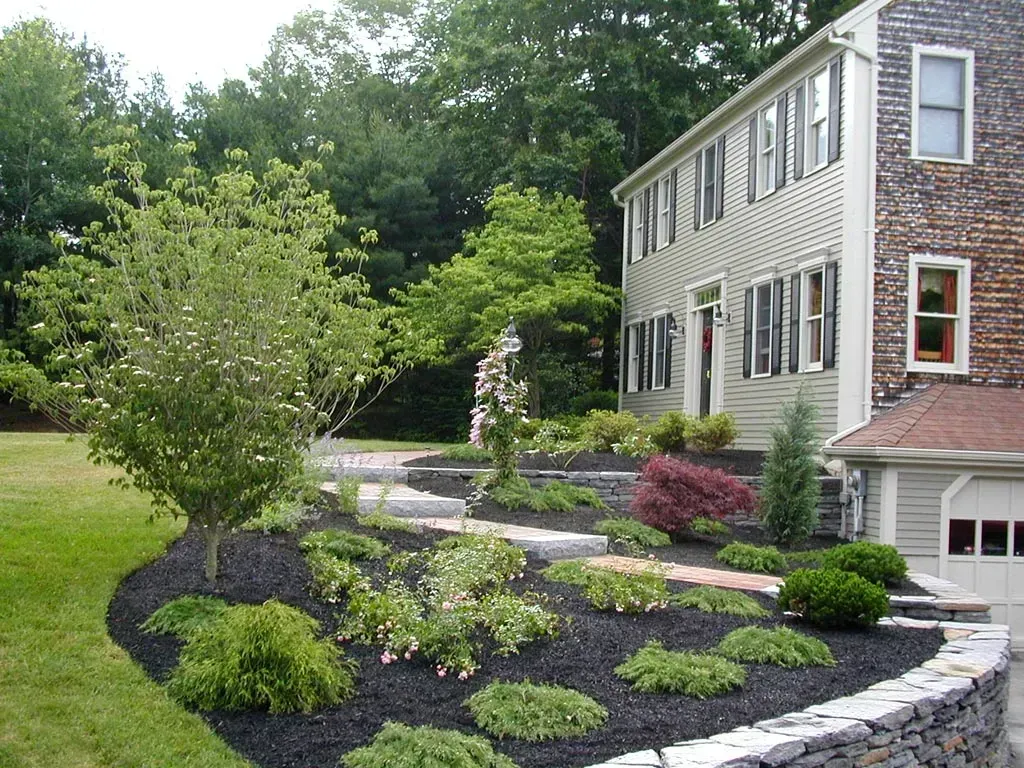 A side view of a two-story light-colored house with a stone retaining wall and landscaped yard filled with mulch and shrubs.