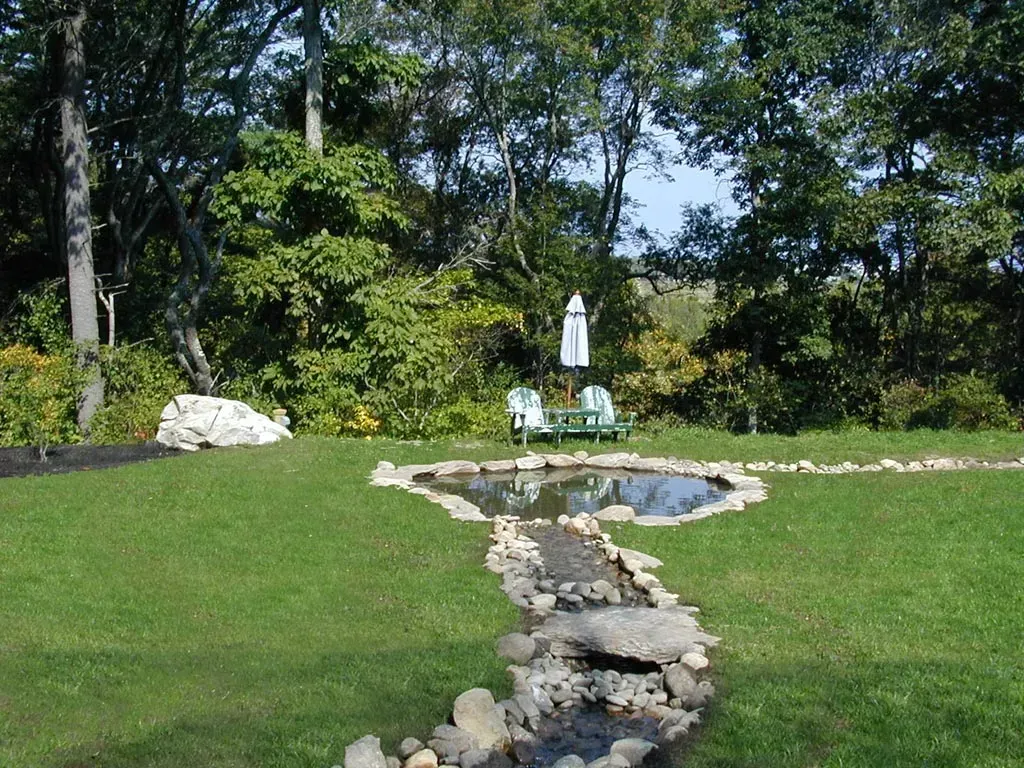 A stone-lined stream leads to a small garden pond in a grassy yard, with Adirondack chairs and an umbrella in the back.