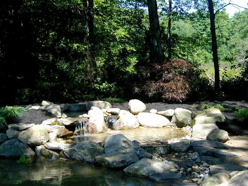 A small, stone-lined garden waterfall flows into a tranquil pond surrounded by lush trees and greenery.