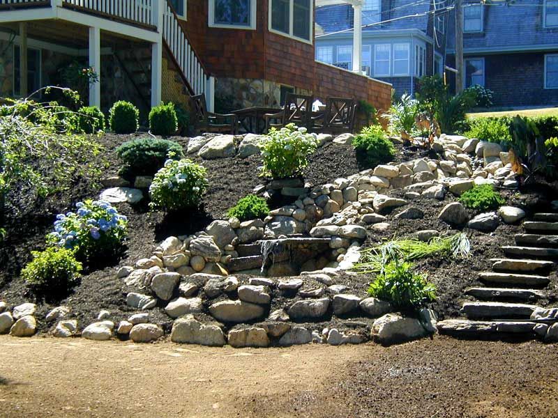 Landscaped sloped yard featuring a rock garden with a small waterfall, stone stairs, and lush green shrubs.