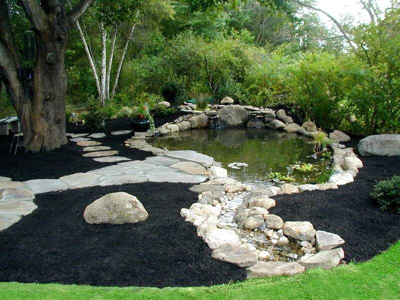 A garden pond with a rocky waterfall, surrounded by dark mulch, stone pathways, and lush green trees.