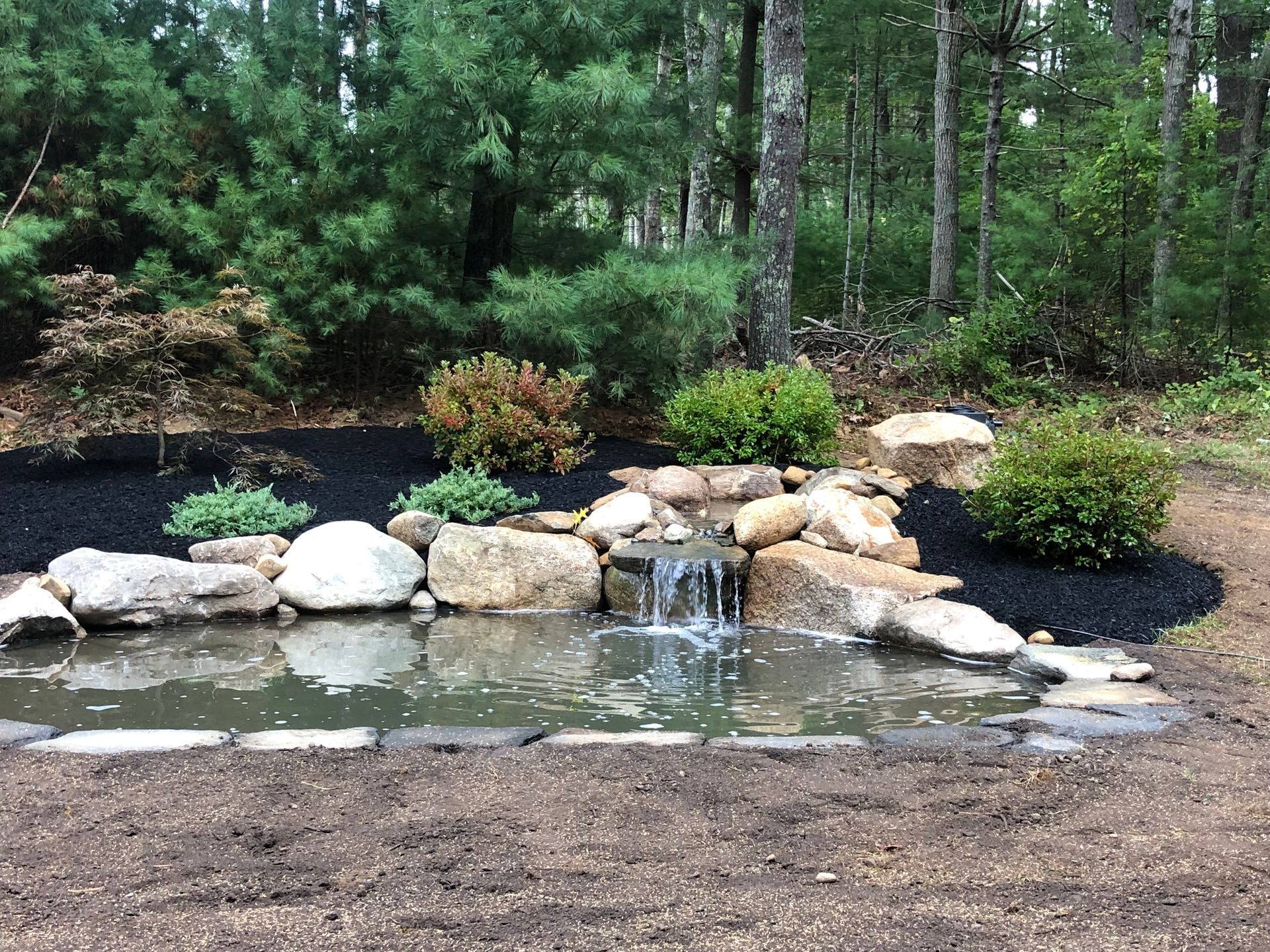 A small garden waterfall cascades over rocks into a pond surrounded by mulch and greenery in a wooded setting.