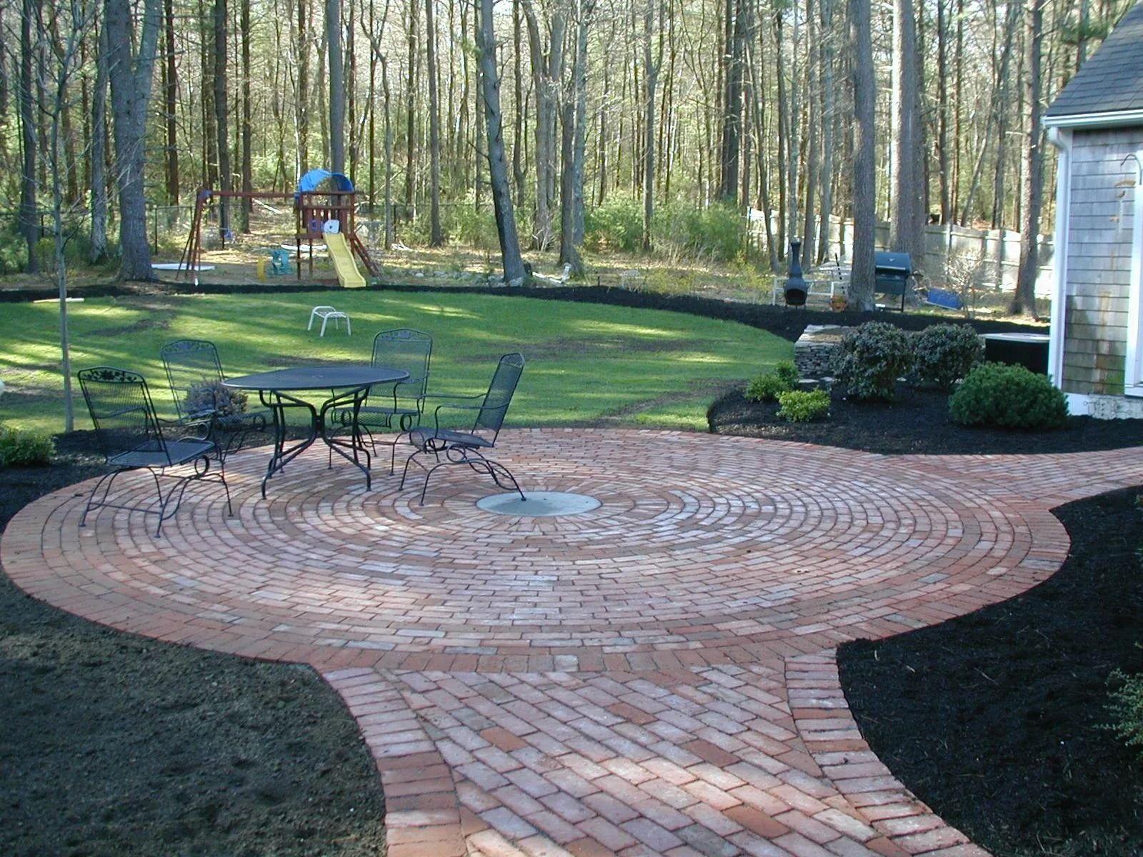 A circular brick patio with a dining set, surrounded by a grassy lawn, mulch, and trees in a backyard setting.