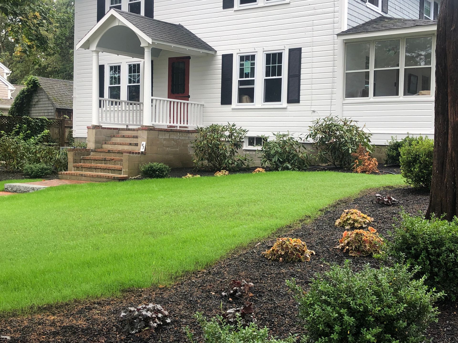 A two-story white house with a covered porch and front stairs overlooks a landscaped lawn with dark mulch beds.
