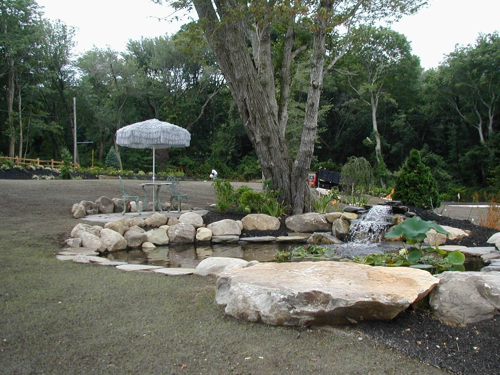 A small stone pond with a waterfall feature and a nearby patio umbrella set in a grassy yard surrounded by trees.
