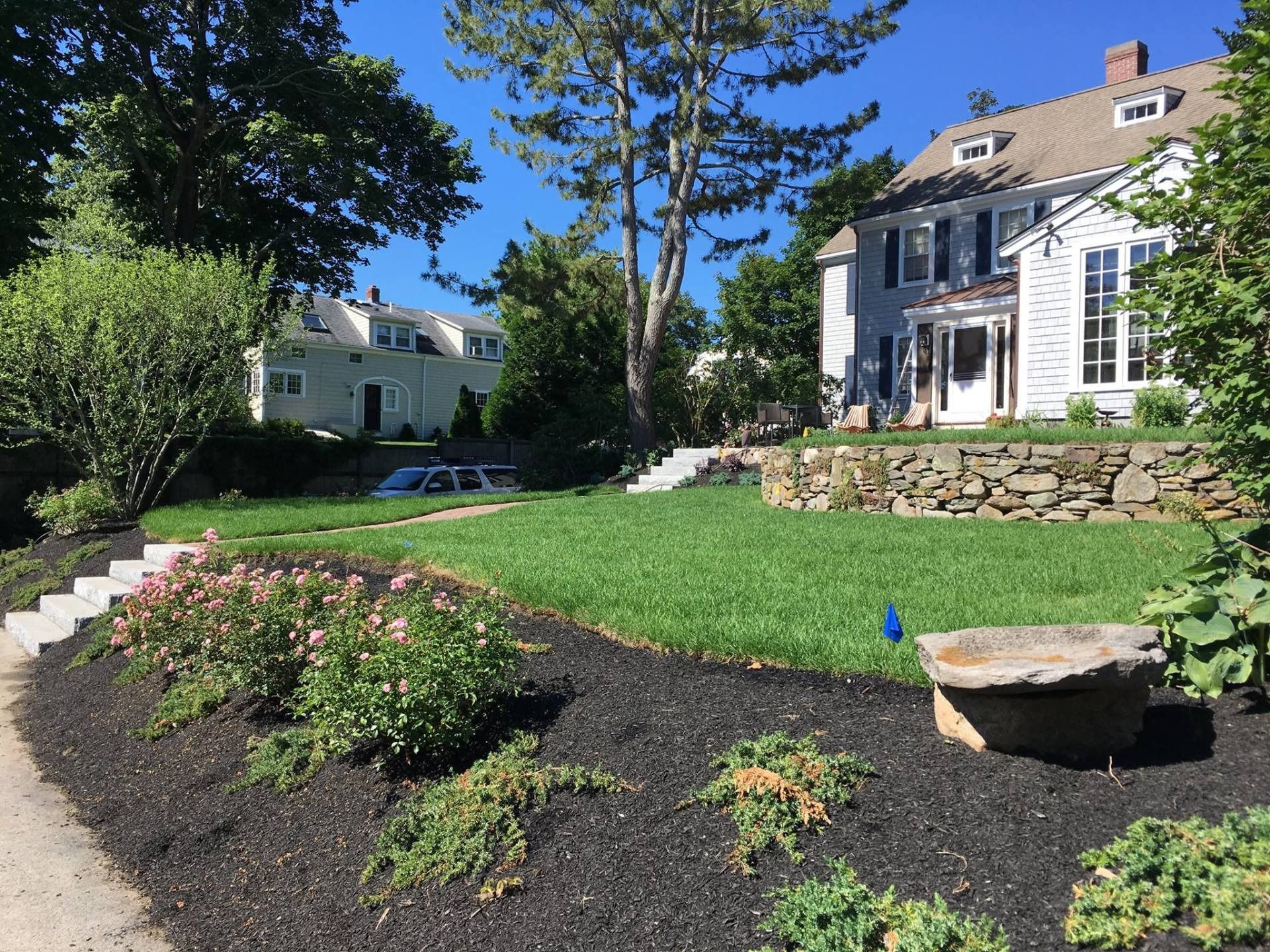 A sunny front yard with a stone retaining wall, green lawn, mulched garden beds, and two white houses in the background.
