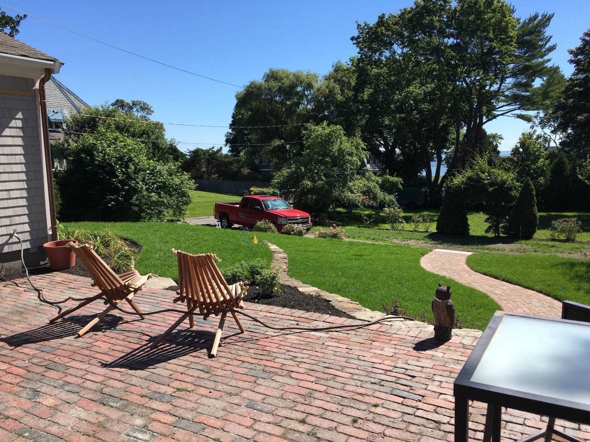 Brick patio with two wooden chairs and a table overlooking a grassy lawn with a red truck and trees in the background.