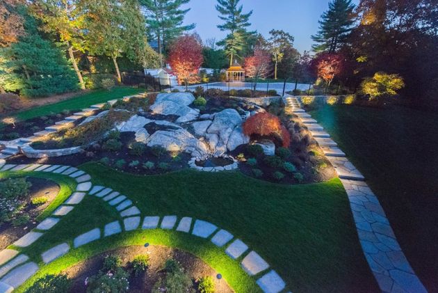 An aerial view of a garden at dusk, featuring illuminated stone pathways winding through lawns, rock gardens, and trees.