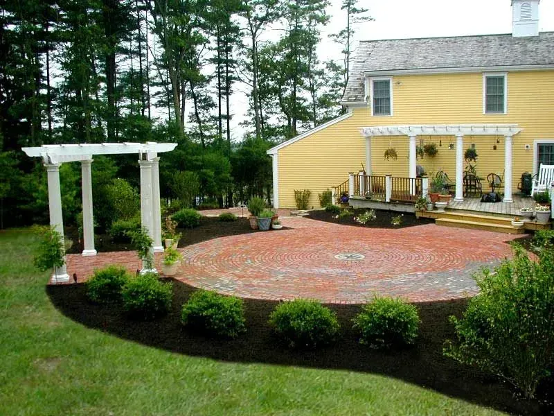 A yellow house with a circular brick patio, pergola, and landscaped garden beds on a sunny day.
