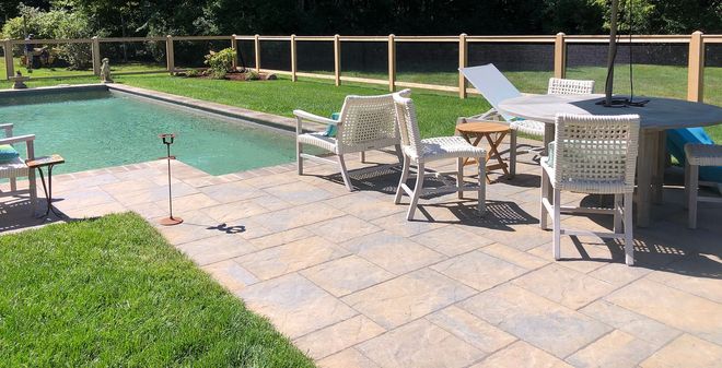 A worker in a cap and gloves kneels on a gravel surface, laying red paving bricks to form a patio.