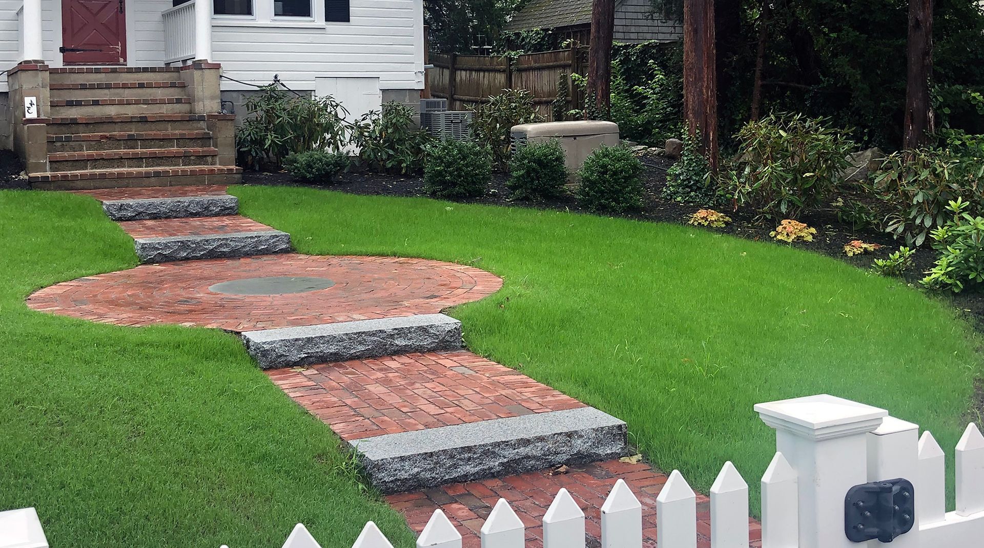 A paved patio with a circular pattern, outdoor dining furniture, and decorative stone planters in a grassy backyard.