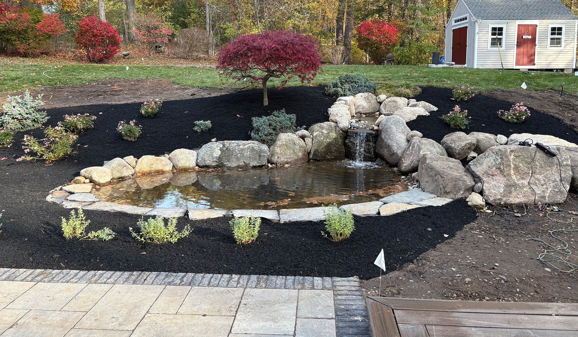 A sunny garden features a stone fountain flowing into a pond with two small green islands, surrounded by blooming flowers.