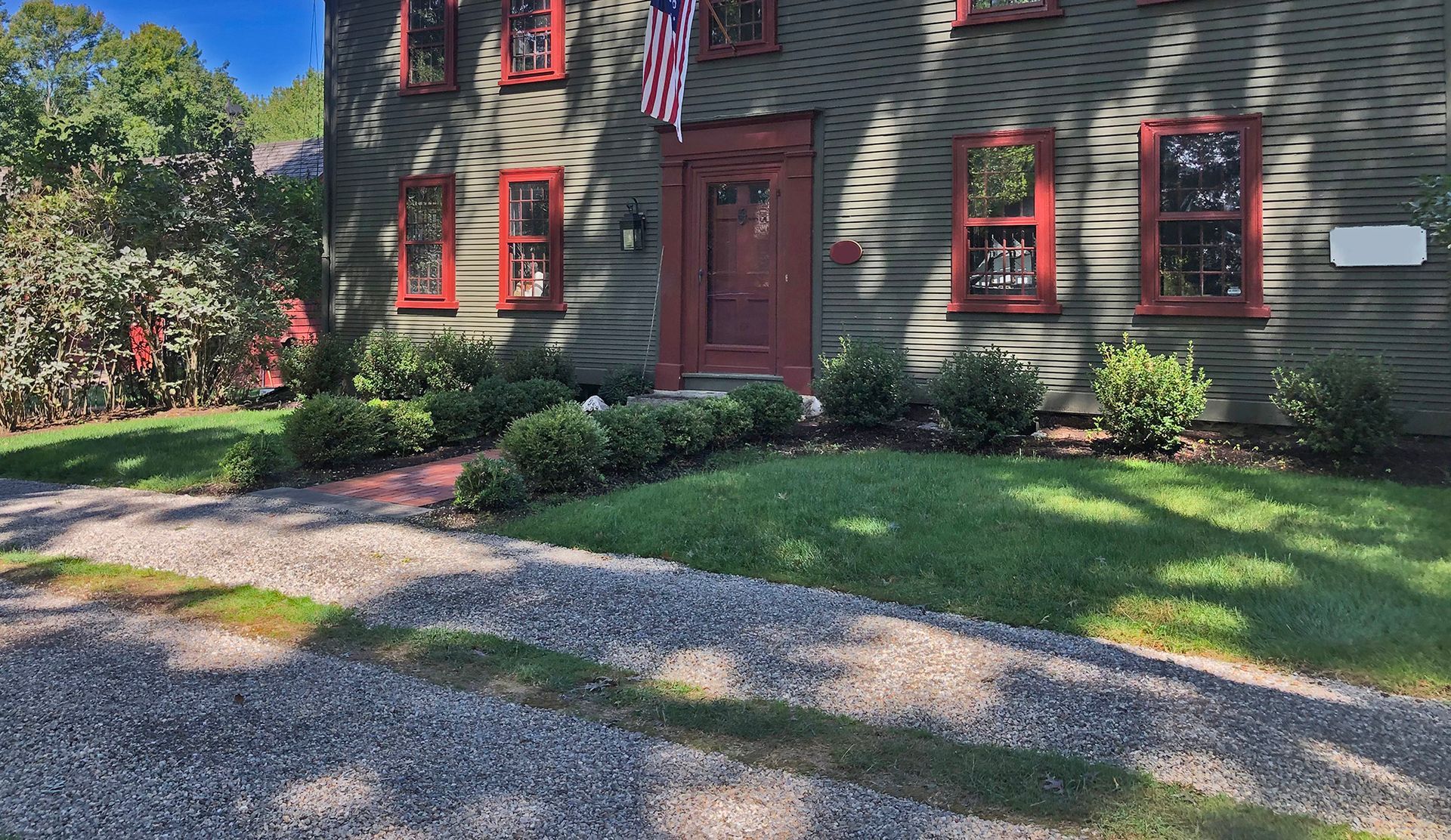 A person wearing a plaid shirt and gloves uses a gas-powered hedge trimmer to shape a green hedge outdoors.