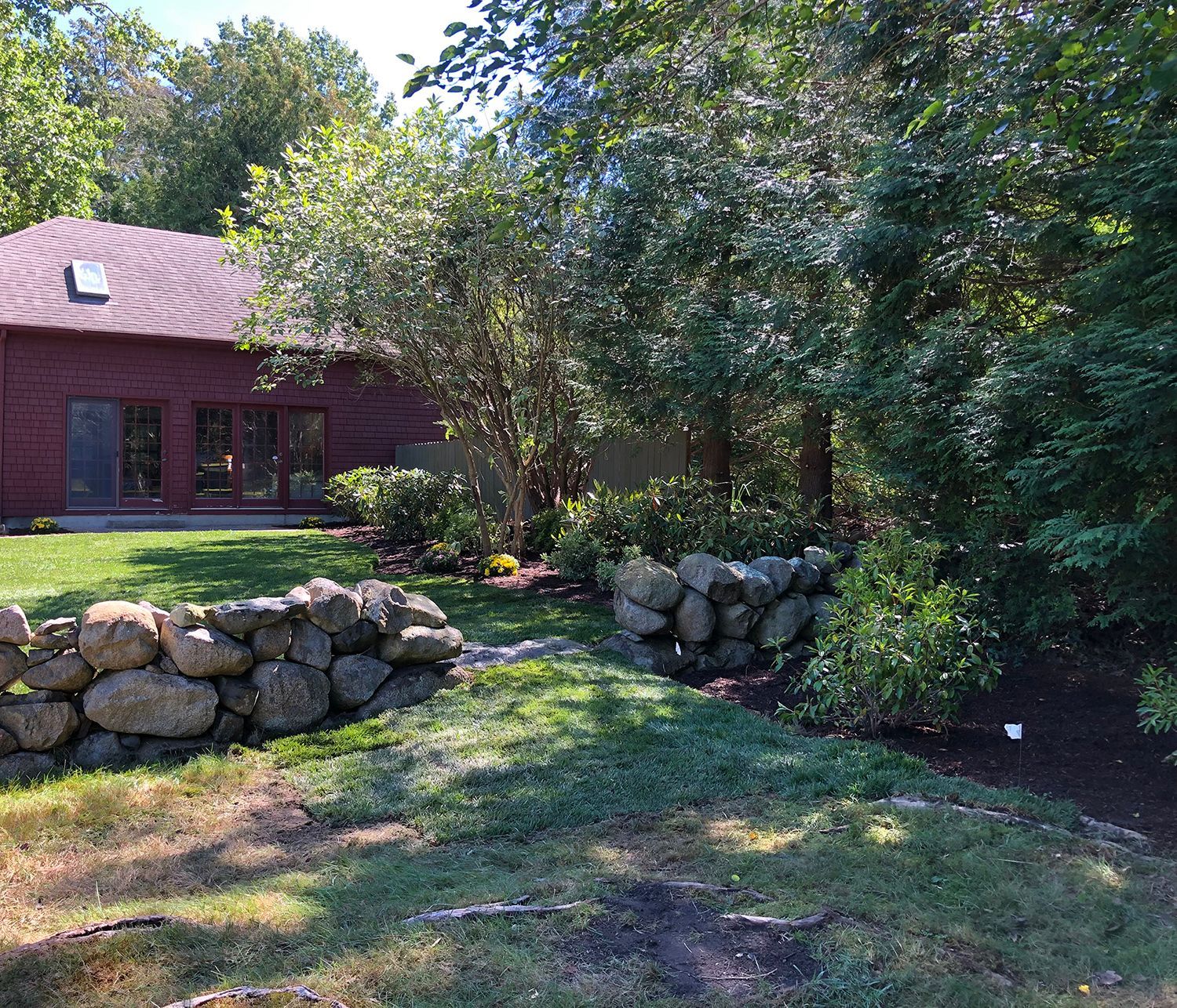 A freshly mowed green lawn stretches toward a lush, sunny garden bordered by varied evergreen and deciduous trees.