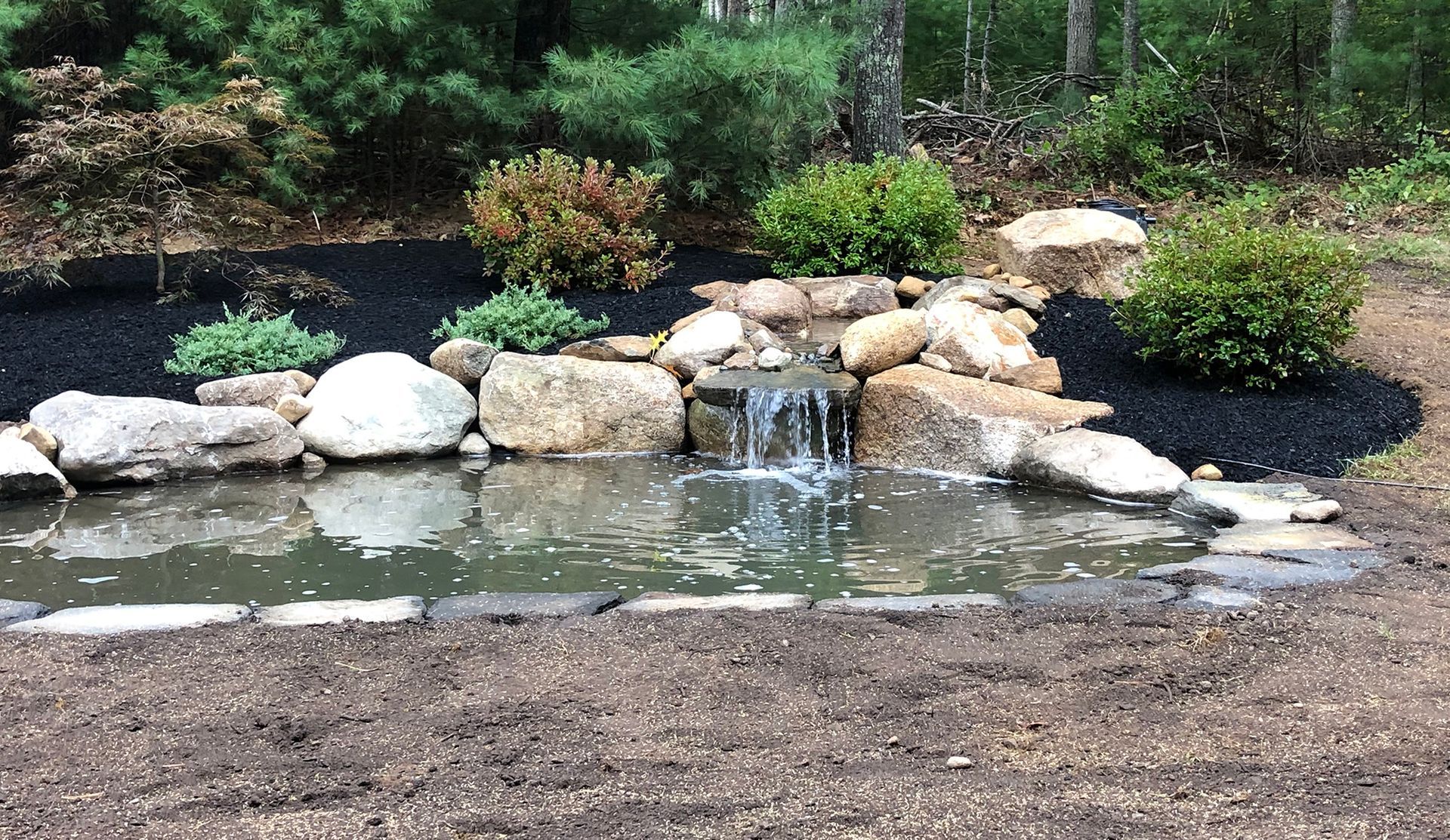 A man-made stone waterfall flows into a pond at sunset, surrounded by pine trees and lush greenery.