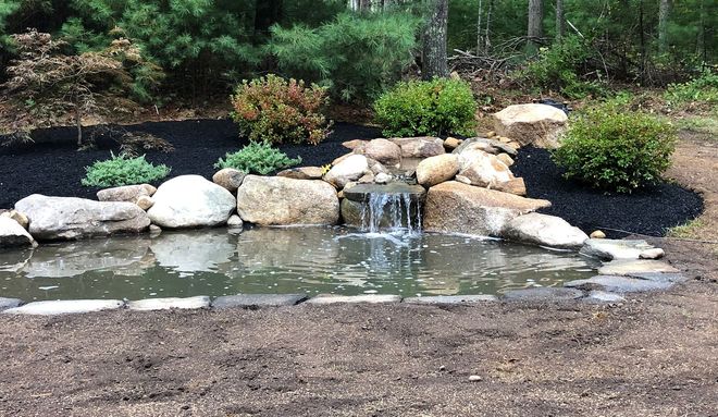 A man-made stone waterfall flows into a pond at sunset, surrounded by pine trees and lush greenery.