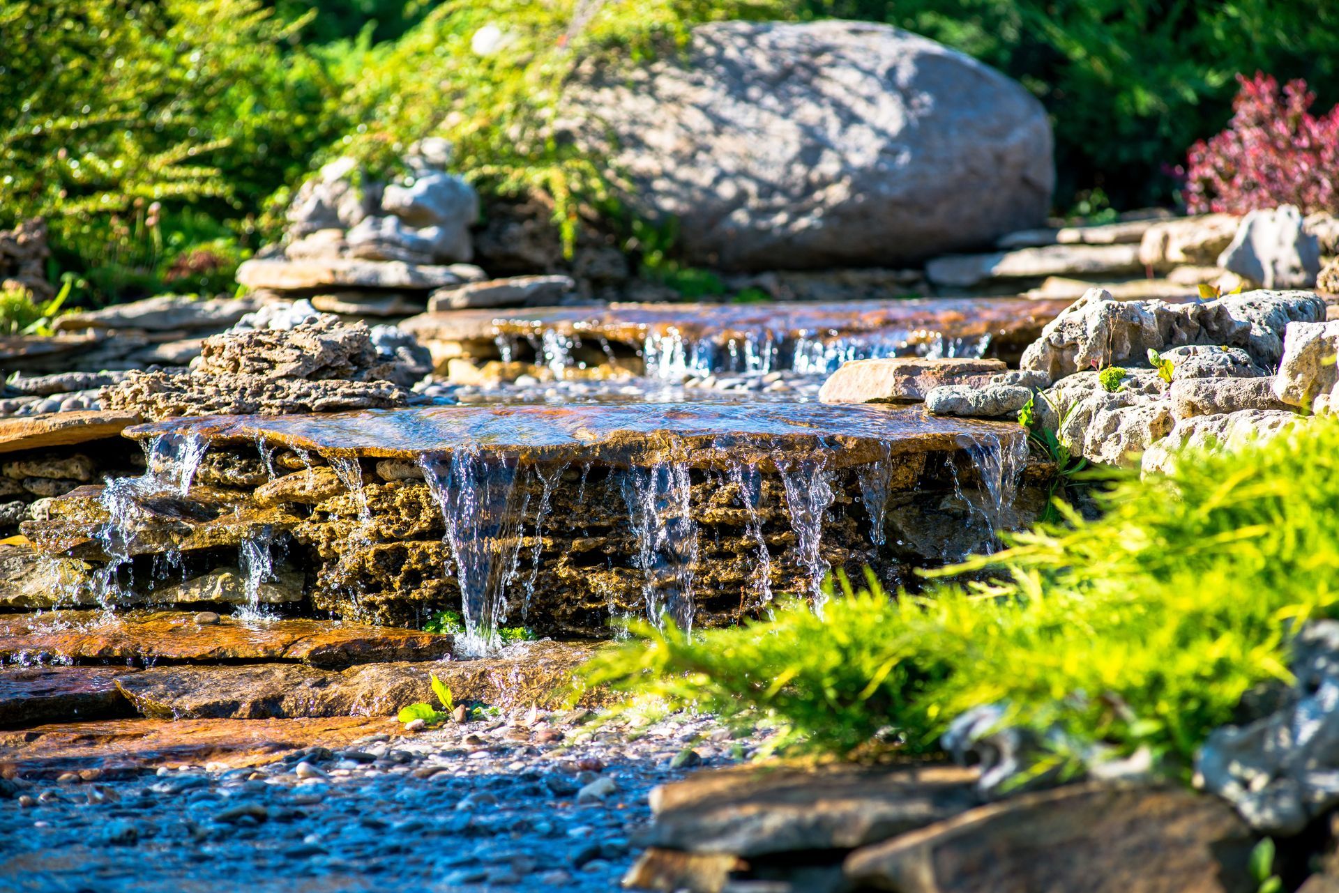 A cascading garden waterfall flows over tiered, mossy stones surrounded by green foliage and a large rock in the background.