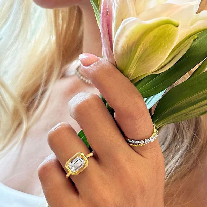 Womans hand with two rings holds white and yellow flowers. One ring has a rectangular gemstone.