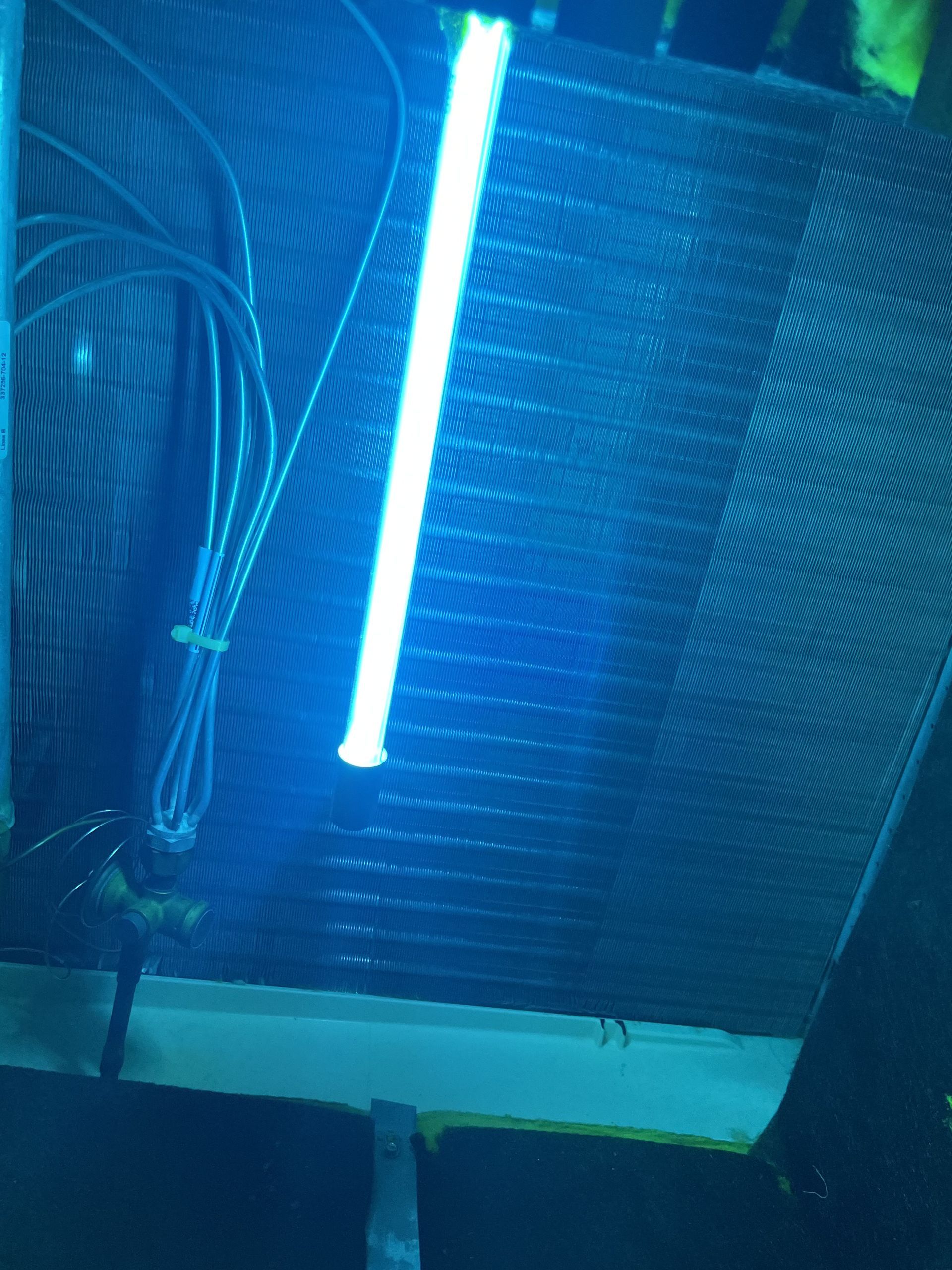 Blue UV light illuminates an air conditioner coil, showing tubes, fins, and surrounding dark components.