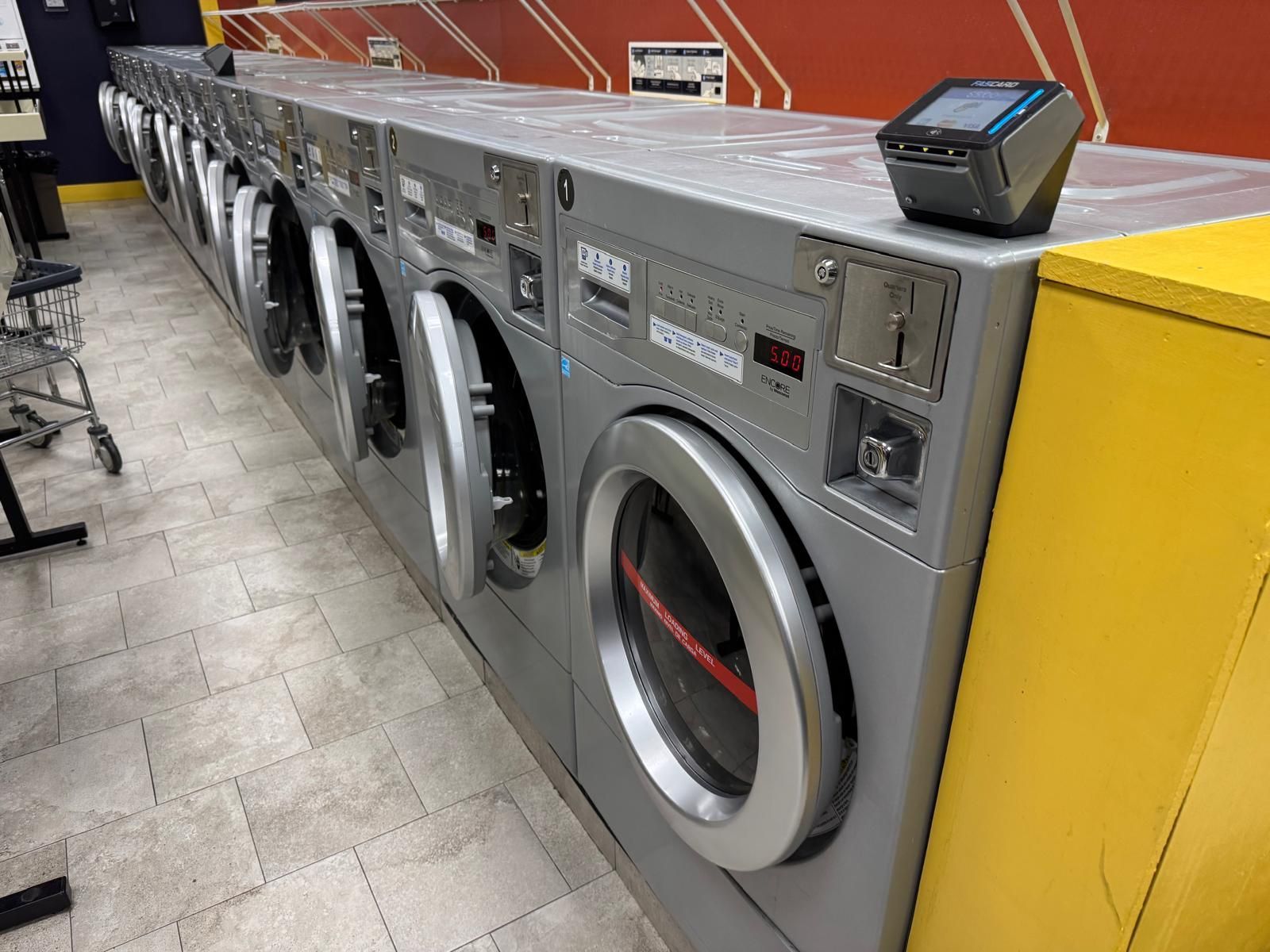 Row of gray washing machines in a laundromat, with a payment device on top of one. Yellow and red walls.