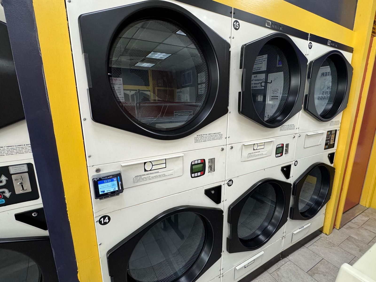 Row of commercial clothes dryers in a laundromat, white with black accents and glass doors.