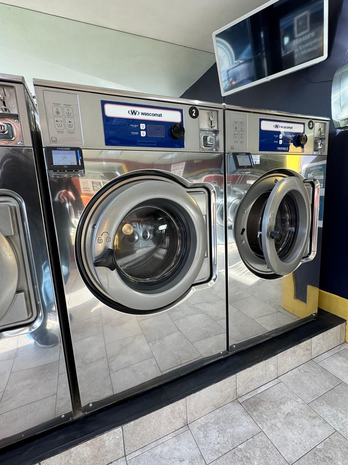 Two stainless steel commercial washing machines in a laundromat. One door open, blue control panels.