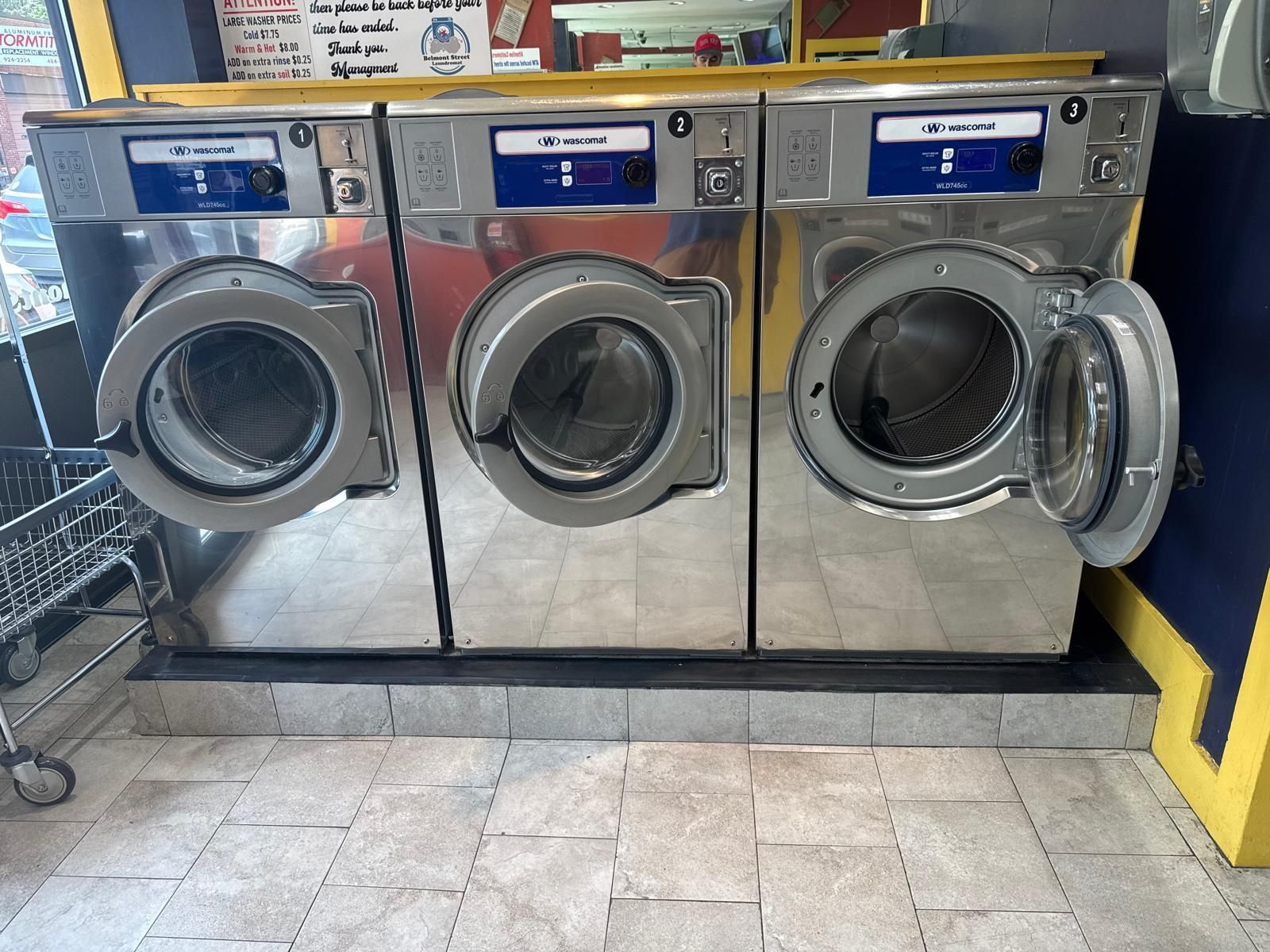 Three industrial washing machines in a laundromat; one door open. Silver with blue panels.