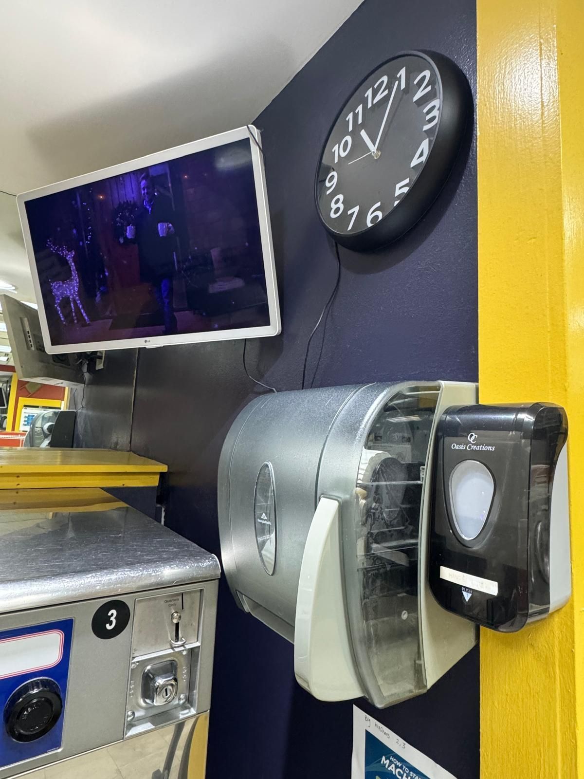 Wall-mounted TV, clock, and soap dispensers on a blue wall next to a yellow pillar. A vending machine is visible.