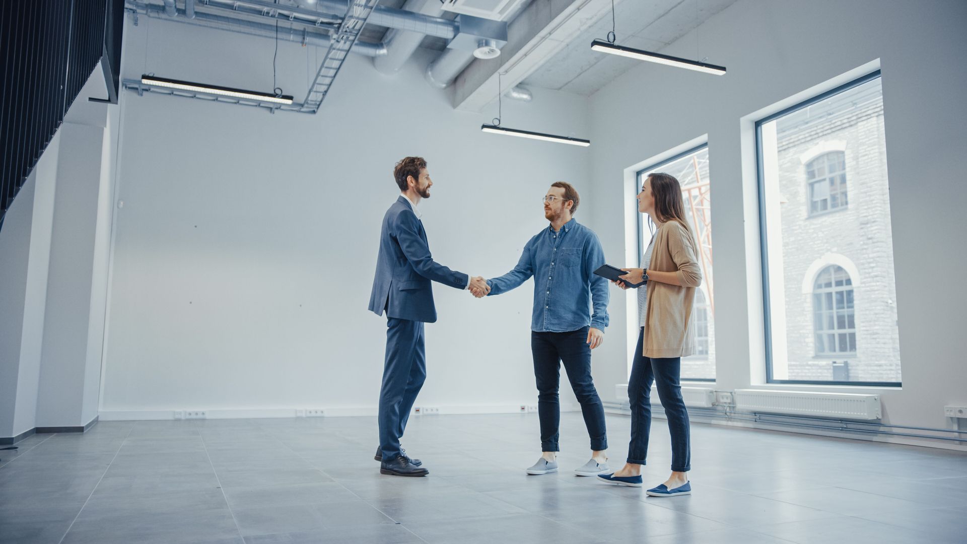 Three people in a bright empty office shake hands near large windows.